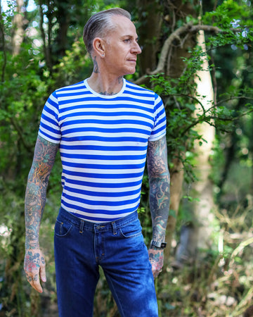 tattooed male model is stood outside amongst trees wearing the Royal & White Stripe Short Sleeve T Shirt with blue jeans. He is posing facing the camera and looking off to one side with one hand in his pocket and other resting by his side.