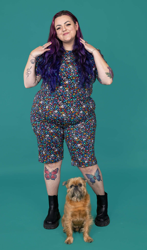 The Boogie Bones Stretch Cycle Shorts worn by a femme alternative tattooed model with long pink and purple hair, the matching tshirt and black boots on a teal studio background with a small brown dog. She is facing forward smiling running her hands through her hair. The black base shorts features dancing smiling skeletons with multicoloured flowers in various sizes and types all over.
