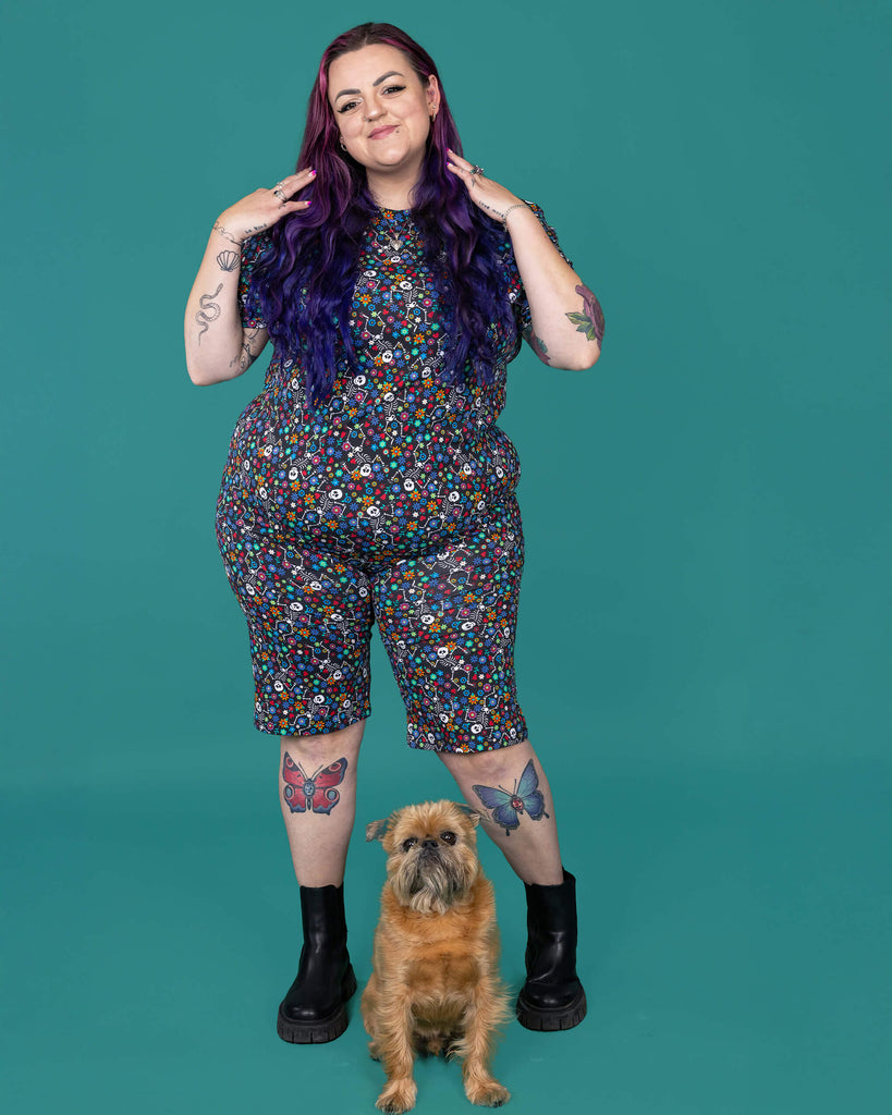 The Boogie Bones Stretch Cycle Shorts worn by a femme alternative tattooed model with long pink and purple hair, the matching tshirt and black boots on a teal studio background with a small brown dog. She is facing forward smiling running her hands through her hair. The black base shorts features dancing smiling skeletons with multicoloured flowers in various sizes and types all over.