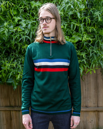 A man with long hair and glasses wearing a dark green Run & Fly striped jumper with a front zip and blue, white, black, and red stripes across the chest. He stands against lush green foliage and a wooden fence. The jumper features ribbed cuffs and hem, reflecting the brand's ethical slow fashion style.