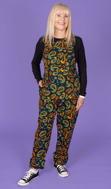 Full-body shot of a model wearing Run & Fly bright paisley dungarees with black and white trainers. The dungarees feature a vibrant paisley design on a black background, with the model standing against a purple backdrop.