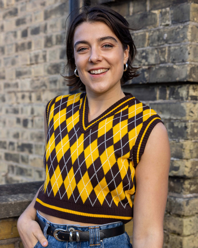 Freya a dark short haired white female model is wearing a brown and gold argyle knitted cropped tank with jeans stood in front of a brick wall