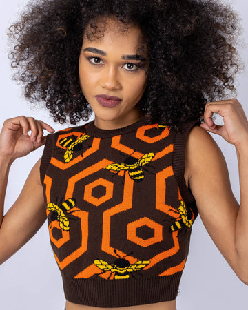 Model is stood in front of a white studio background wearing the brown bee 70's honeycomb knitted tank top with leather look leggings. They are facing the camera with both hands holding onto the top of the tank top.