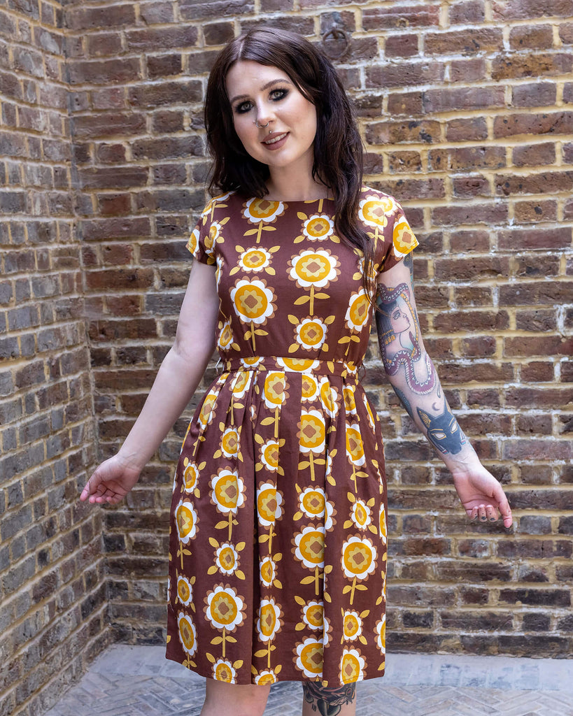 Florence a brown long haired white model with tattoos is wearing a brown retro tea dress with floral print against a brick wall. 