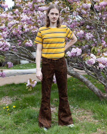 Jack, a male model with long blonde hair is stood outside in East Sussex by a blossom tree wearing Hendrix Tan Paisley Corduroy Retro Bell Bottom Flares paired with Gold Sunset Stripe Retro T Shirt. The trousers are a tan colour with an all over black paisley print. Jack is posing facing the camera with his arms down by his sides.