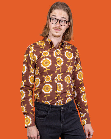 Jack a white male with shoulder length blonde hair and glasses in his 20's wearing a retro brown shirt with long selves and black jeans shot against an orange studio background.