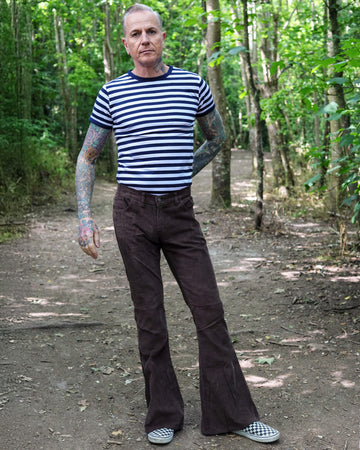 Model is stood in a forest area wearing the brown corduroy bell bottom super flares with a navy and white stripe t-shirt and checkered van shoes. They have one hand in their pocket with one hand behind their back, they are stood facing the camera with one leg slightly out to the side to show off the flares.