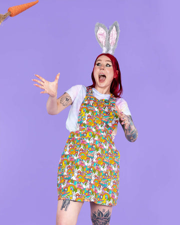 Florence a red haired tattooed femme white model wearing a Waffle Mama Design rainbow bunny print pinafore in multi colours with mushrooms, flowers, rainbows and clouds. She has paired this with a rainbow crop top, grey bunny ears and black trainers. She is facing forward posing on a purple background looking shocked as a carrot is thrown towards her.