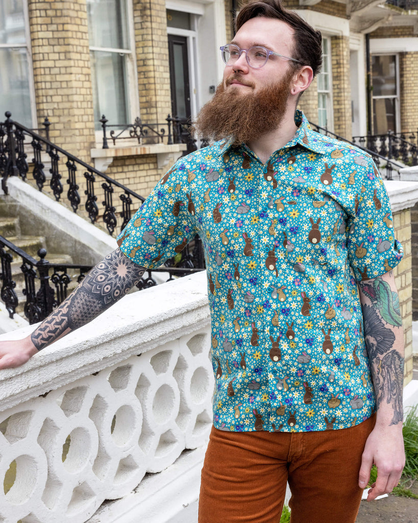 Dec is wearing the Bunny Meadow short sleeve blue shirt with floral pattern, worn with brown corduroy stretch flare trousers. Model is looking away from the camera outside of a street.