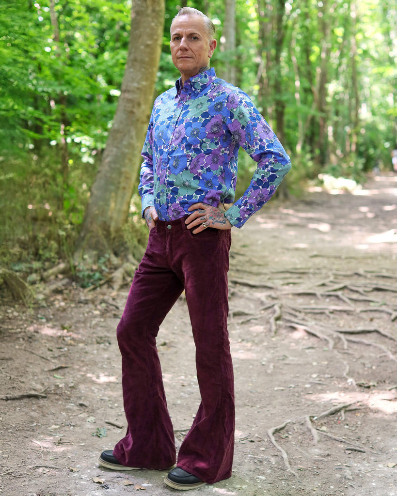 Model is stood outside in a forest area wearing the burgundy corduroy bell bottom super flares with a purple and blue floral shirt and dark brown boat shoes. They are facing slightly to the right and posing with both hands on their hips and one leg angled out to show off the flares. 