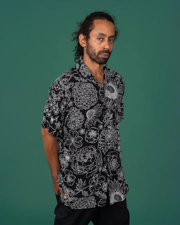 A man with curly hair tied back, wearing a black shirt adorned with intricate white floral designs. He is standing against a plain green background, looking slightly to the side with a neutral expression.