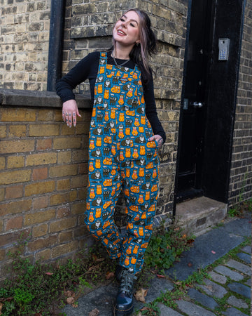 Model standing casually against a brick wall, wearing teal dungarees featuring a whimsical cat and mouse print by Waffle Mama Design, paired with black boots and a black long-sleeve top.