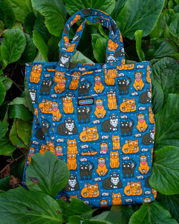 “Cat and Mouse” tote bag displayed on green foliage. The print showcases orange and black cats, each accompanied by tiny mice on a deep blue background, highlighting the quirky, nature-inspired aesthetic of the design.