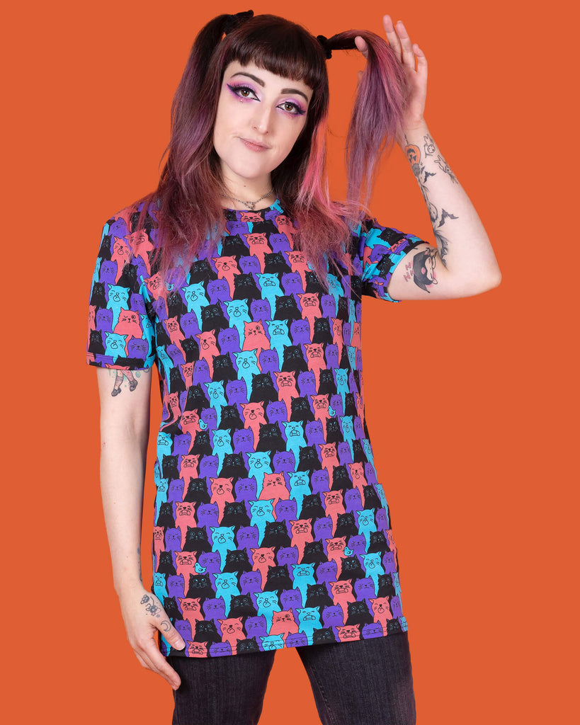 Sarah is stood in front of an orange background wearing the Cat Chorus Short Sleeve Tee with black stonewash denim flares. She is facing forward with one hand flicking her hair and the other hand resting by her side. The cat chorus print is an all over repeating pattern of sky blue, peachy red, purple and black cats pulling various faces and a sporadic sky blue bird.