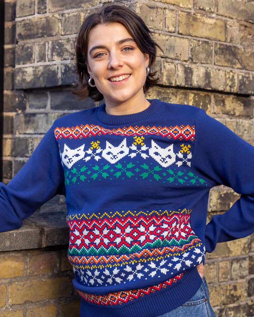 Freya is a white woman in her 20's with short brown hair wearing a blue christmas style jumper with cats on. She is leaning on a brick wall