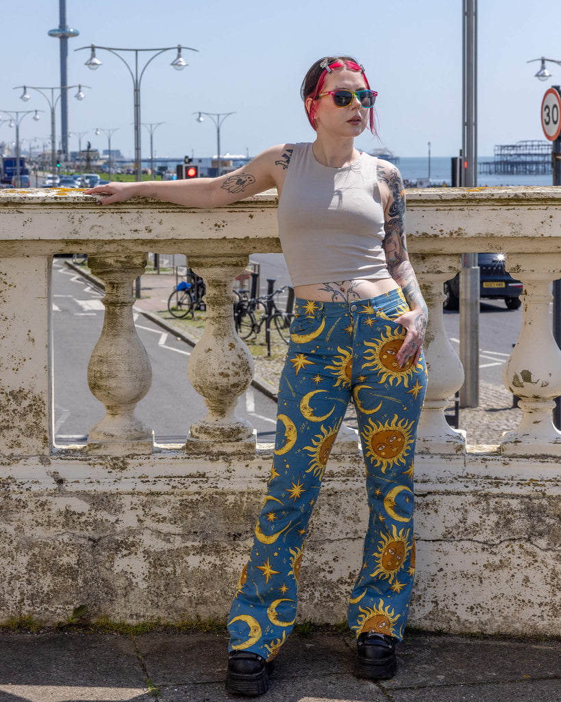 The Celestial Sun and Moon High Waisted Bell Bottom Flares shown on a femme tattooed model with red and black hair and a beige tank top, rainbow sunglasses and black trainers. She is stood outside in front of Hove seafront. She is facing forward leaning on a wall with one hand in the flare top pocket smiling looking over to one side. Print is gold 90s style moons and suns with faces, and sparkles all on a blue background.