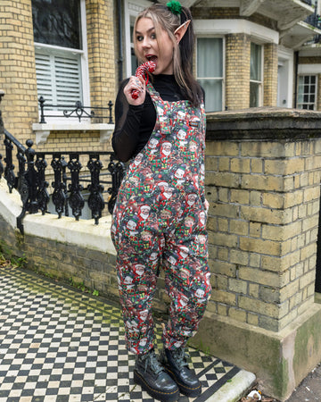 Full-length shot of a model in Run & Fly Christmas-themed dungarees, paired with black mesh sleeves and glittery boots. The design showcases Santa, reindeer, and other festive prints, creating a whimsical holiday fashion statement. Photographed outdoors with Victorian architecture in the background.