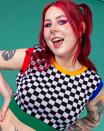 Flo wearing the Run and Fly Colour Block Checkerboard Cropped Knitted Tank Top on a teal background. She has styled the all over black and white checkerboard vest with a yellow trim neck, red left arm trim, blue right arm trim and green hem trim with oversized y2k cargo trousers, silver hoop earrings and neon pink eye makeup. Flo has red hair in bunches with piercings and tattoos. She is facing forward leaning in smiling with both hands on her hips.