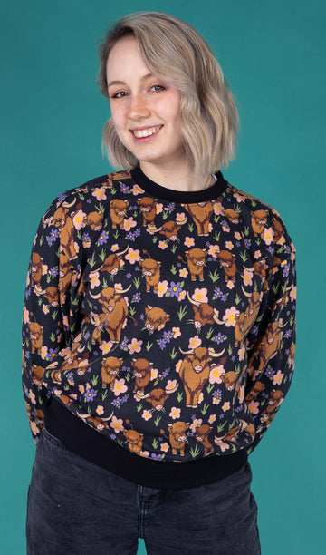 The Highland Cow Sweatshirt worn by a femme model with short blonde hair with dark denim jeans on a teal studio background. She is facing forward smiling to camera with both hands resting behind her. The black base sweatshirt has pink, yellow and purple flowers, grass bunches and various brown highland cows all over with a black neck, hem and sleeve trim.