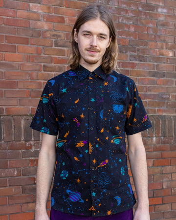 Jack is stood in front of a brick wall wearing the cosmic space short sleeve shirt with purple trousers. They are facing forward posing with both arms resting by their side and smiling to camera. Photo is cropped from the hips up.