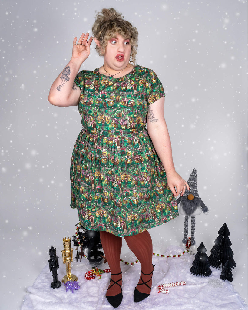 Maddie strikes an amused pose, placing one hand behind their ear while wearing the cosy cottage print dress. Snowfall adds to the winter wonderland atmosphere, while Maddie’s brown tights and black shoes complement the green-toned dress, decorated with detailed illustrations of whimsical cottages.