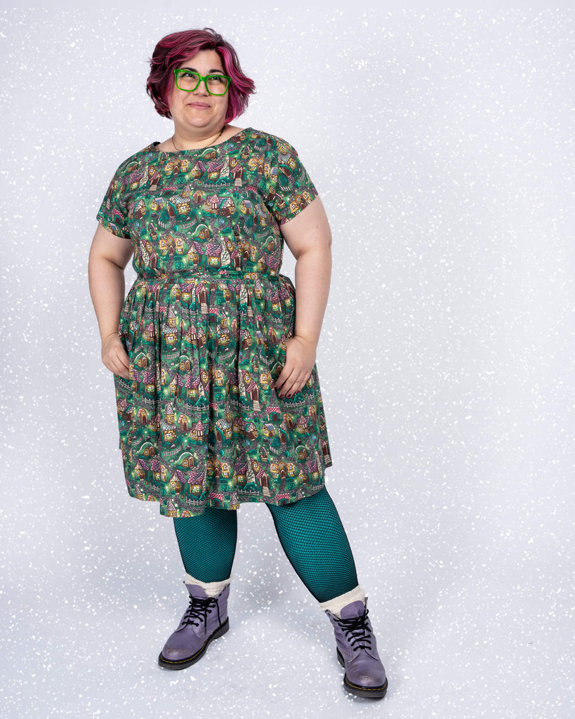 Full-body shot of Jackie in the “Cosy Cottage” tea dress by Lauren Ash Design. The fitted bodice and enchanting cottage-themed pattern create a playful and comfortable look, styled with teal tights and purple boots.