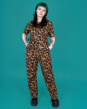 Faeryn, a tattooed model with green and black hair is wearing a black jumpsuit with various sizes of brown highland cows on it, some eating grass. There is also various peach, purple and yellow flowers on the design. The jumpsuit is paired with black boots. The model is posing with their hands in the jumpsuit in front of a green background. 