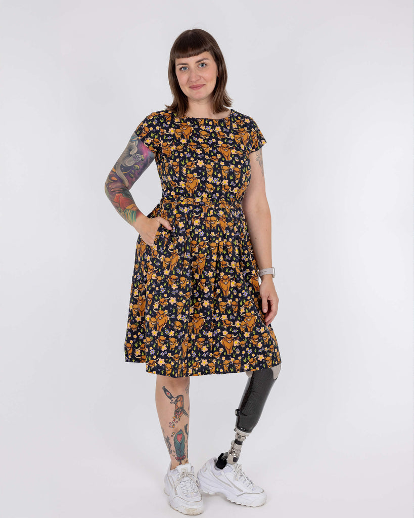 Laura is wearing a highland cow printed tea dress in front of a white background in a studio in Hove. She has a prosthetic leg and is smiling at camera with her hand in the dress pocket