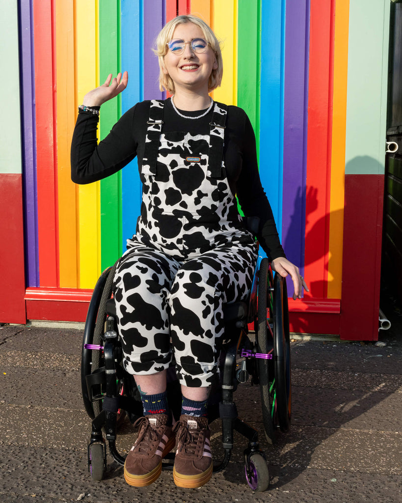 A non-binary model in a wheelchair wears Run & Fly Cow Print Stretch Twill Dungarees with a black top. They pose with a cheerful smile in front of a vibrant rainbow wall. The dungarees, made by a small ethical slow fashion brand, feature a bold cow print pattern, adding a playful and stylish touch.