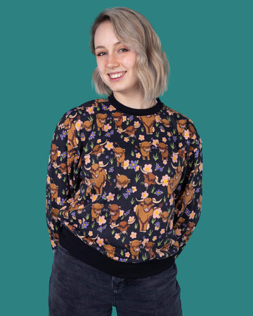 The Highland Cow Sweatshirt worn by a femme model with short blonde hair with dark denim jeans on a teal studio background. She is facing forward smiling to camera with both hands resting behind her. The black base sweatshirt has pink, yellow and purple flowers, grass bunches and various brown highland cows all over with a black neck, hem and sleeve trim.