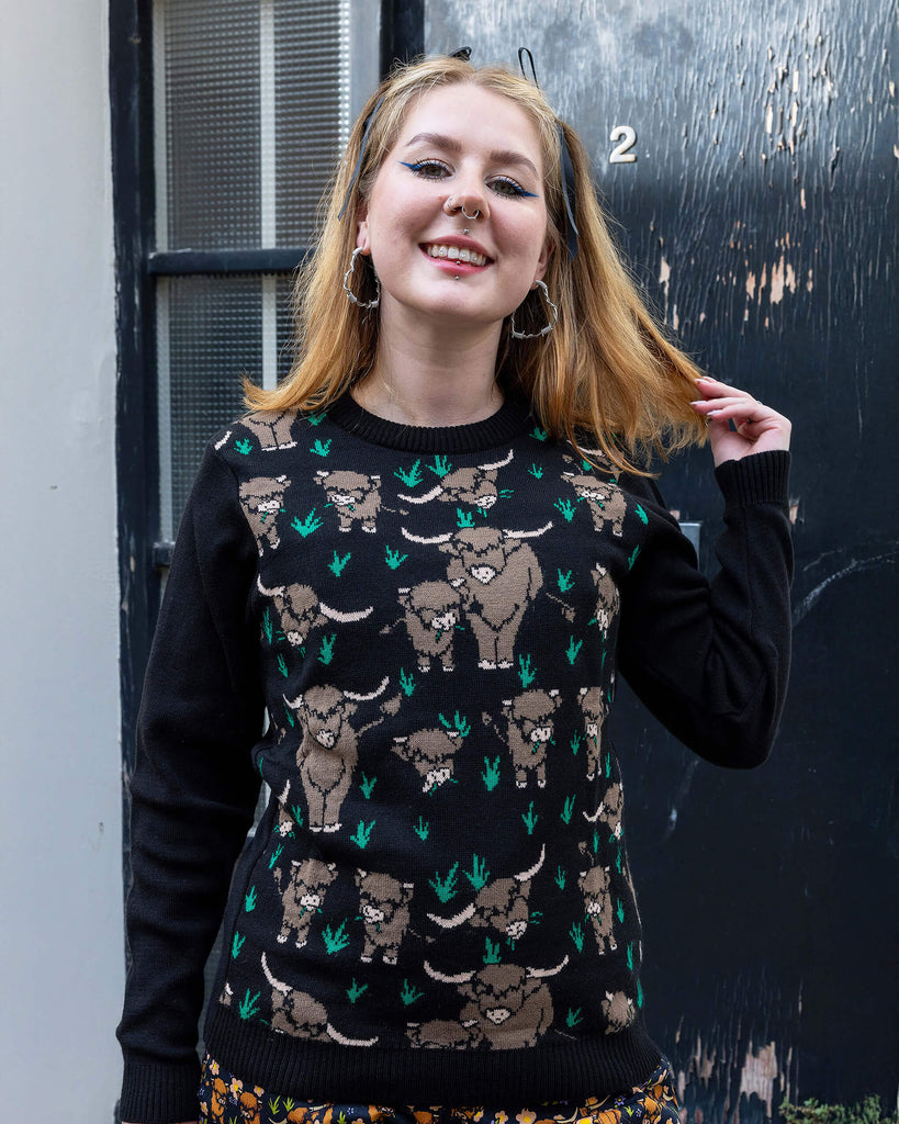 Florence, a femme model with blonde hair is stood outside in Hove wearing Highland Cow Jumper on top of Highland Cow Dungarees. The jumper is black with highland cows on the torso. Florence is smiling facing the camera with one hand in her hair. 