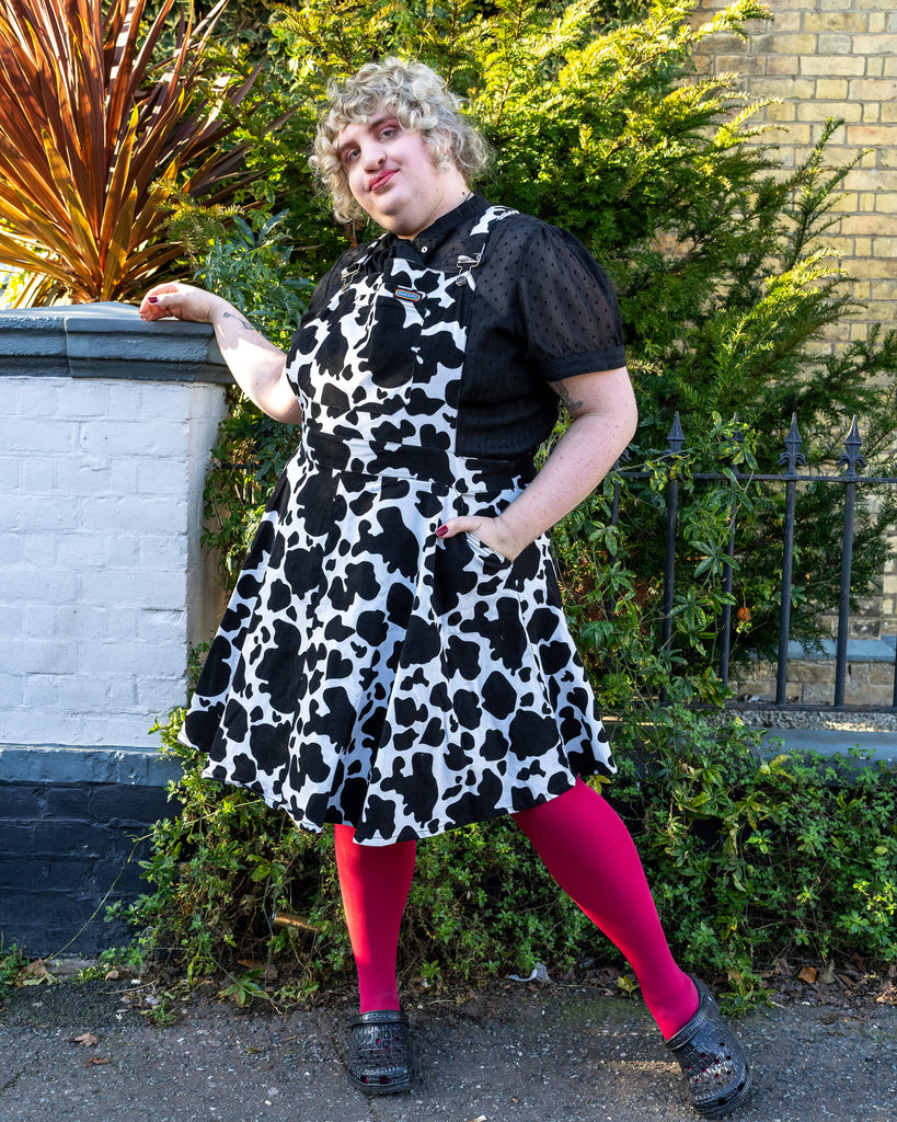 Full Outfit with Cow Print Pinafore - Maddie poses confidently in the Run & Fly cow print pinafore dress, styled with pink tights and a black blouse. The cow print pattern adds a quirky, bold look to the outfit, complemented by her expressive pose.