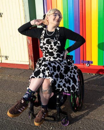 Image shows Eliza, a non-binary model in a wheelchair, wearing Run & Fly’s cow print flared pinafore dress against a colourful rainbow backdrop. The black and white cow print dress has a flattering A-line shape, paired with a black long-sleeve top and chunky trainers. Eliza's blonde hair is styled with a polka dot bow. Ethical slow fashion by Run & Fly.