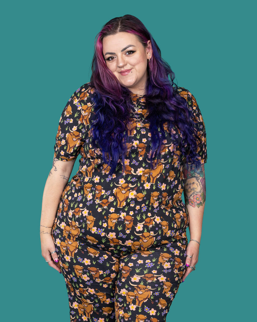 The Highland Cows Short Sleeve T Shirt worn by a femme alternative tattooed model with long pink and purple hair, the matching cycle shorts on a teal studio background. She is facing forward smiling with her arms resting by her side. The black base t shirt features grass, purple and yellow flowers and brown highland cows eating grass.