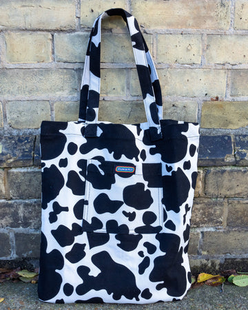 A Run & Fly cow print tote bag with bold black-and-white spots, featuring a front pocket adorned with the rainbow Run & Fly label. The bag is propped against a rustic brick wall.