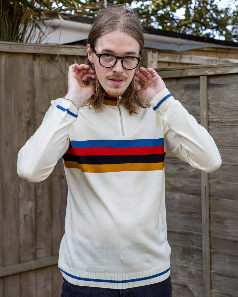 Jack a white male with shoulder length blonde hair and glasses is wearing a high neck knit jumpers with stripes across the middle. He has his hands up adjusting his hair and is stood in a garden.