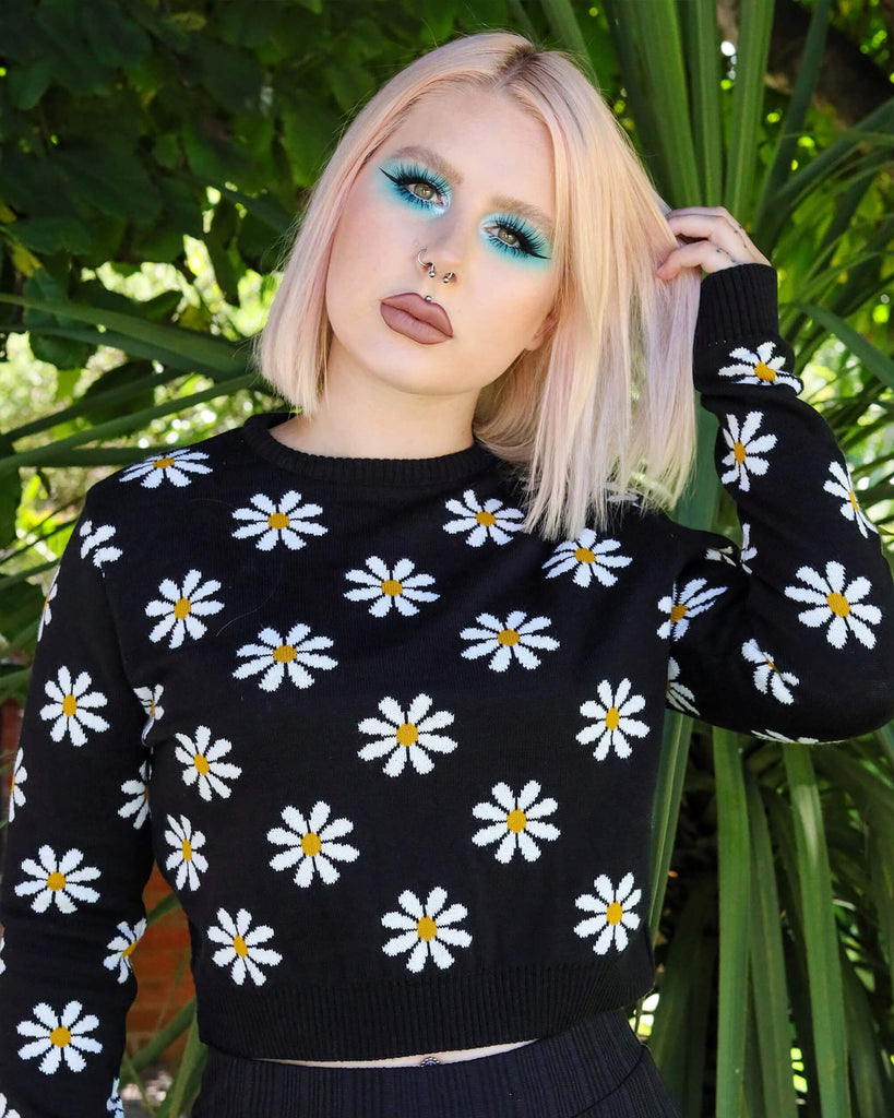 Model is stood in a green garden area wearing the daisy chain black cropped jumper with black trousers. They are facing the camera with one hand in their hair and the other by their side. Photo is cropped from the hips up.