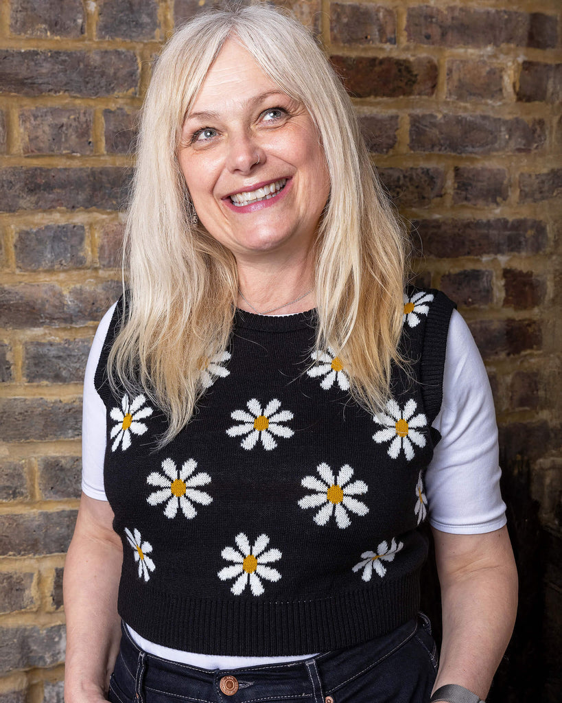 karina a white woman in her 50's has blonde hair and is wearing a daisy knitted tank top and jeans with a white tee against a brick wall