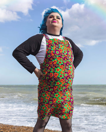 Jeff is stood on a beach wearing the 70s dark floral print stretch corduroy long pinafore dress with a long sleeve black tshirt, tights and white trainers. They are facing forward posing with both hands on their hips and smiling looking up.