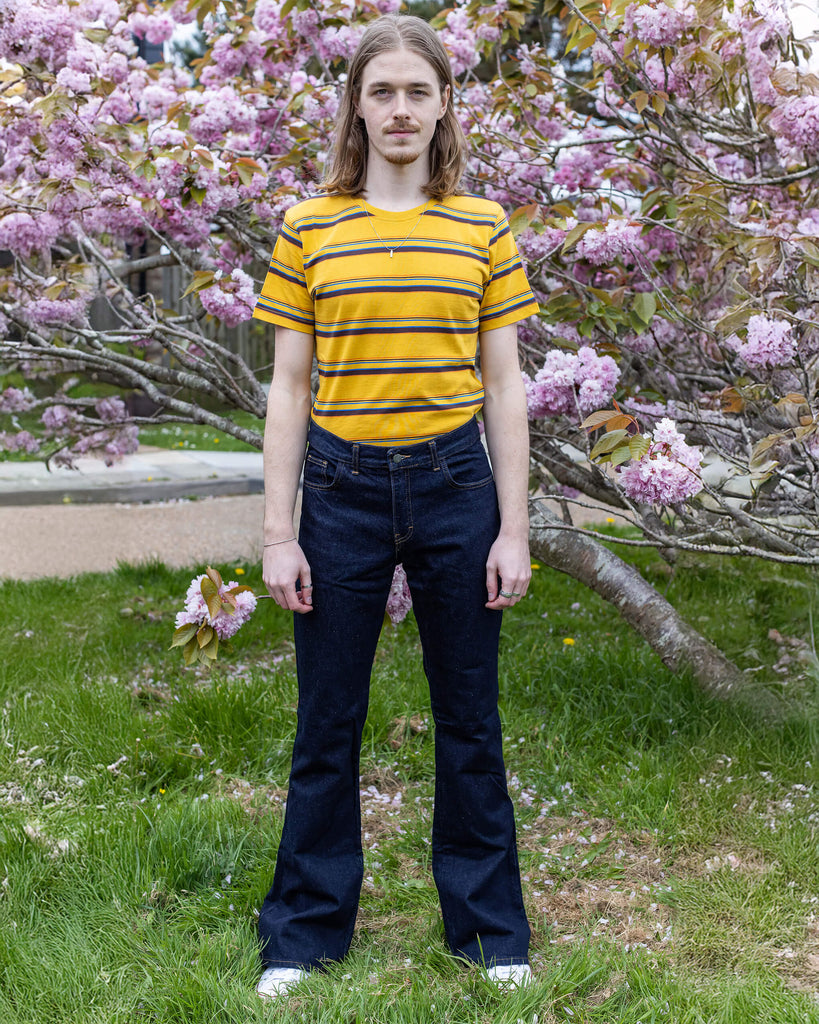 Jack a blonde male model with shoulder length hair is looking at camera infant of a blossom tree wearing a mustard striped tee and indigo blue flares. 