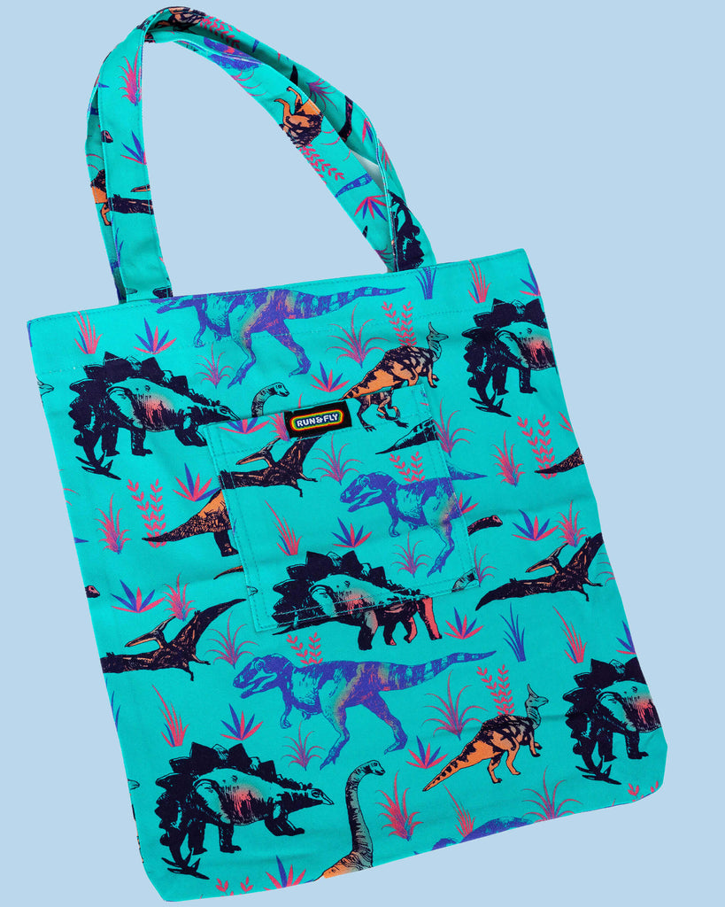 Flat lay of the Jade Adventure Dinosaur Tote Bag on a light blue backdrop. The green tote bag has an all over dinosaur print with a front pocket with rainbow Run&Fly logo on the front.