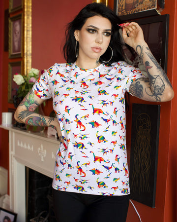  Rosie is stood in front of a red wall containing framed prints wearing the white rainbow dinosaur tshirt with black trousers. They are facing forward posing with one hand on their hip whilst the other running through their hair. Photo is cropped from the hips up.