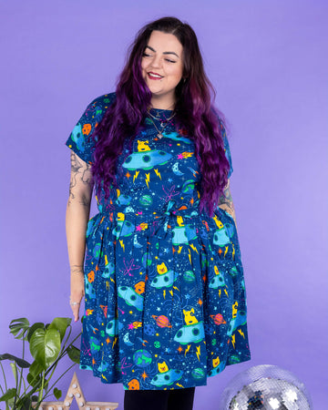 Luisa, a femme model with purple hair and tattoos, is stood in a photography studio in Hove in front of a purple backdrop wearing Dogs in Space Stretch Belted Tea Dress with Pockets with black tights and boots. The print features orange and yellow bull terriers, pomeranians and labradors in blue spaceships on a space themed blue background with stars, planets and rockets. The dress is short sleeved with pockets and a belt tie. Luisa is facing the camera and looking off to one side smiling.