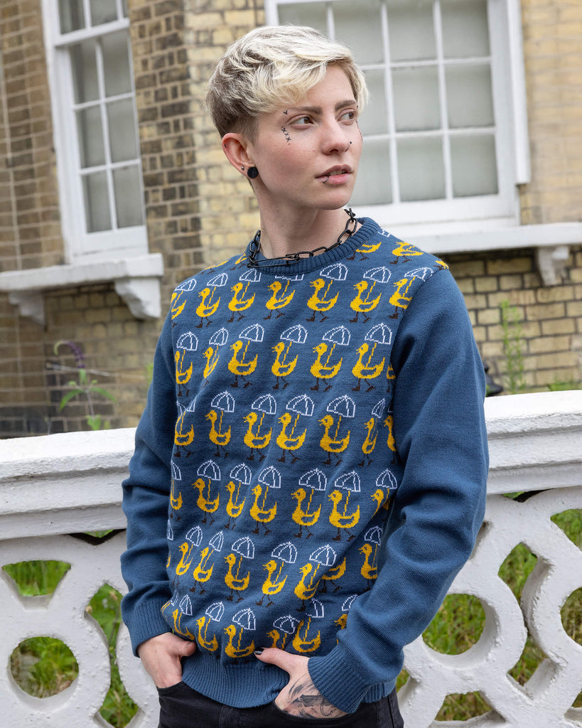 A model wears the Nice Weather for Ducks Jumper by Run & Fly, a small ethical slow fashion company. The blue jumper features a quirky pattern of yellow ducks and white umbrellas, perfect for rainy days and adding a fun touch to any outfit. The model stands outdoors against a white wall with an expressive look, embodying the jumper's playful spirit. Ideal for sustainable fashion lovers seeking unique, cosy knits.