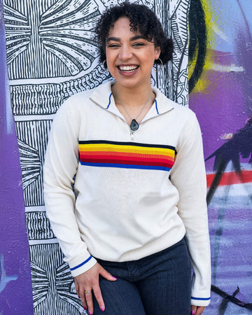 Georgia is stood in front of a graffiti wall wearing the ecru retro zip neck jumper with denim jeans. They are facing forward posing with one hand on their leg and the other hand resting by their side whilst laughing to camera. Photo is cropped from the thighs up.