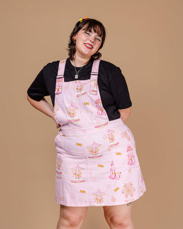 A cheerful model poses in Run & Fly’s Pink Fairground Stretch Twill Pinafore Dress, featuring nostalgic fairground prints of rides and tickets. This ethical slow fashion piece, designed in soft pink, brings retro amusement park vibes to a modern pinafore style. Paired with a black T-shirt, the outfit is a playful nod to childhood memories.