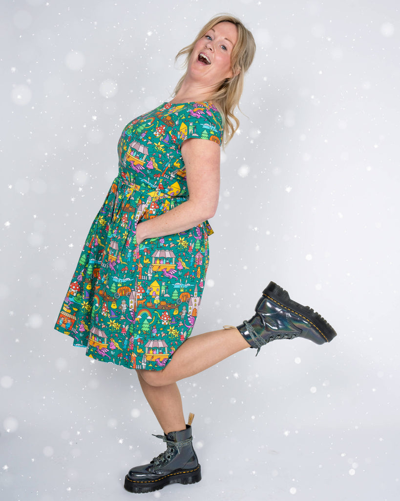 A playful side pose of a model wearing a green tea dress with a fairytale-inspired print. The dress showcases colourful illustrations of cottages, mythical creatures, and magical elements, with the model kicking one leg back and smiling joyfully.