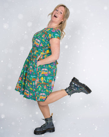 A playful side pose of a model wearing a green tea dress with a fairytale-inspired print. The dress showcases colourful illustrations of cottages, mythical creatures, and magical elements, with the model kicking one leg back and smiling joyfully.