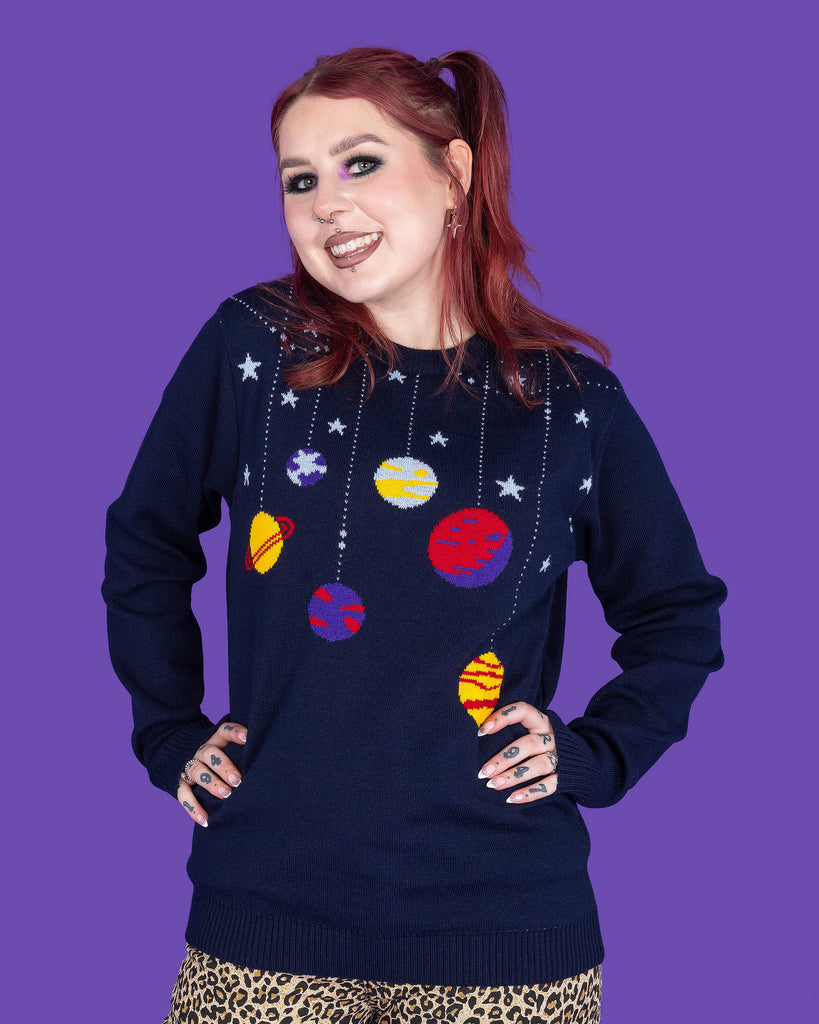 model with red hair and purple eye makeup wearing a navy coloured knit jumper with light blue stars and various planets on it, paired with leopard print jeans. The model is smiling at the camera with her hands on her hips. The background of the photo is purple.