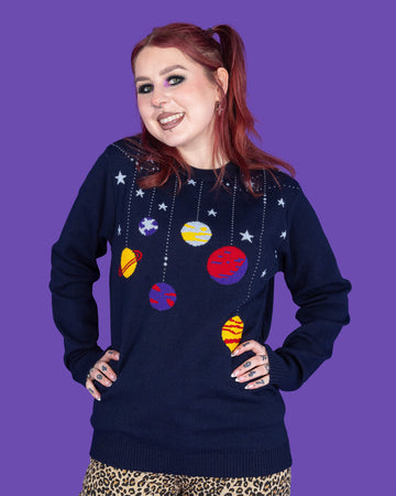 model with red hair and purple eye makeup wearing a navy coloured knit jumper with light blue stars and various planets on it, paired with leopard print jeans. The model is smiling at the camera with her hands on her hips. The background of the photo is purple.